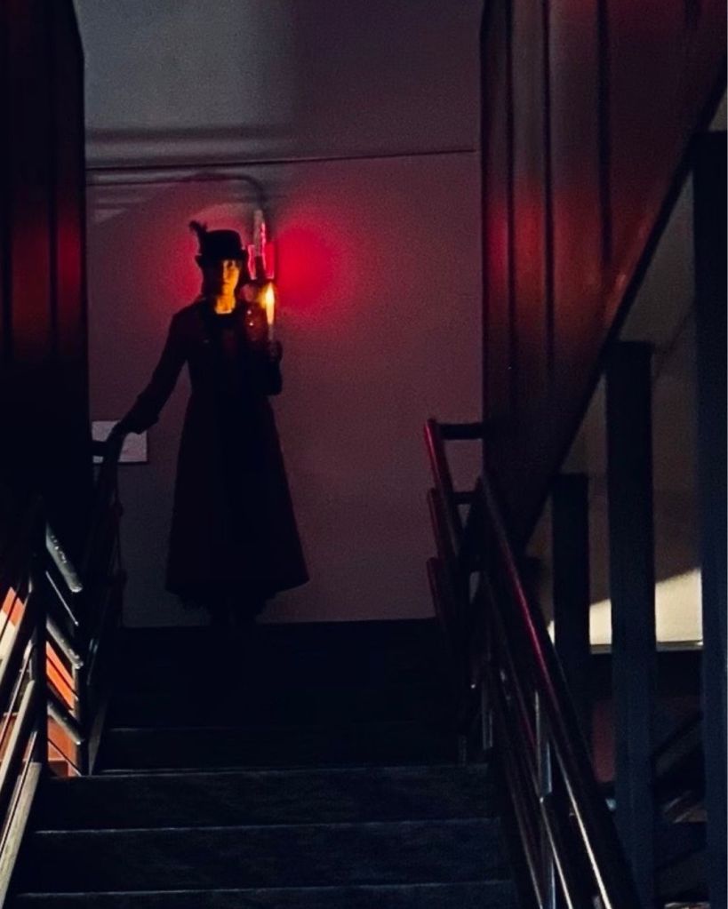 A woman dressed in old-fashioned clothing holds a candle at the top of library stairs
